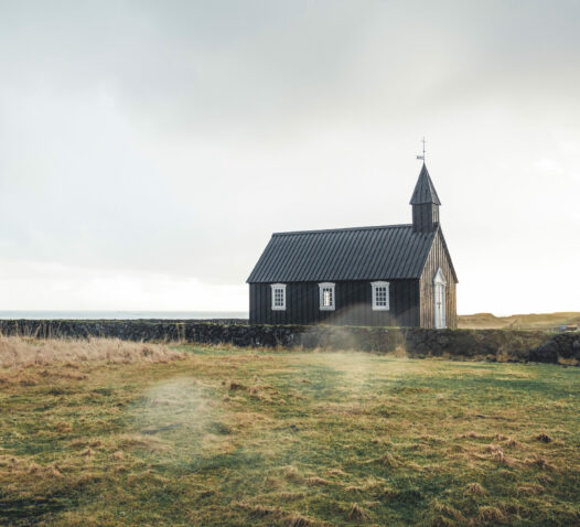 En liten svart kyrka står på en öppen äng under molnig himmel.