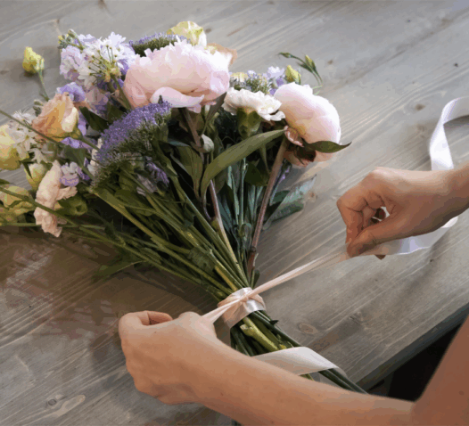En person binder en bukett med blommor med ett band på ett träbord.