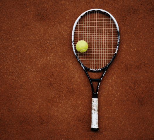 Tennisracket och gul tennisboll ligger på en grusplan.